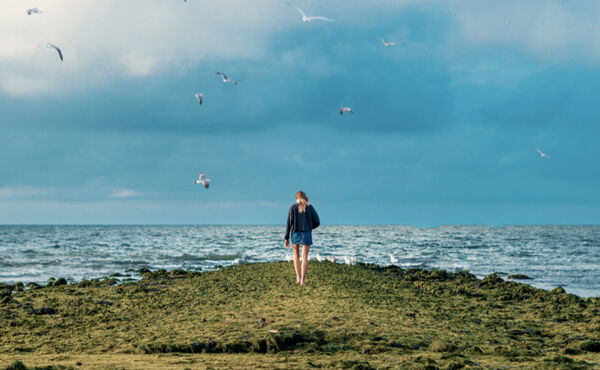 Woman walking alone toward the sea under a cloudy sky. A dreamlike moment symbolising reflection and the subconscious.